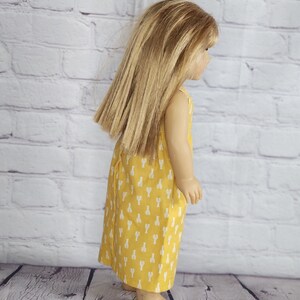 May include: A doll wearing a yellow dress with a white lobster pattern. The dress is sleeveless and falls to the doll's ankles. The doll has long, straight blonde hair and is standing in front of a white brick wall.