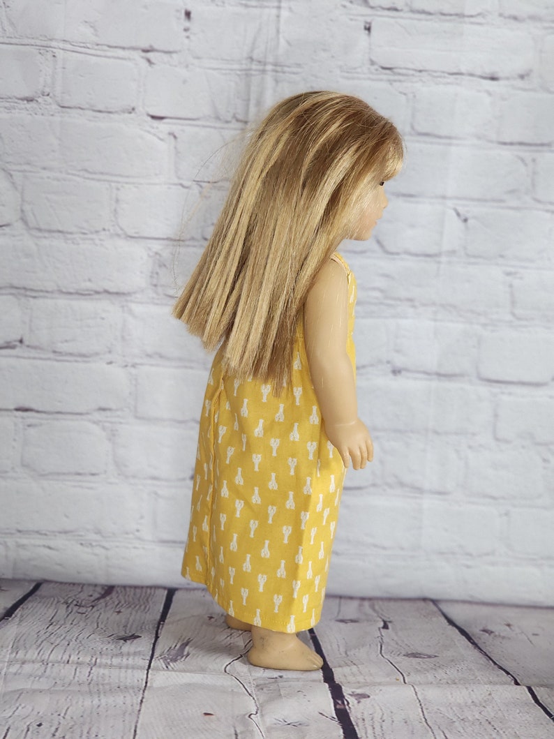 May include: A doll wearing a yellow dress with a white lobster pattern. The dress is sleeveless and falls to the doll's ankles. The doll has long, straight blonde hair and is standing in front of a white brick wall.
