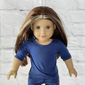 May include: A doll with long brown hair with blonde highlights and blue eyes. The doll is wearing a navy blue shirt and pants. The doll is standing in front of a white brick wall.