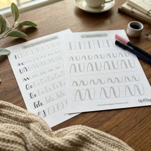May include: Two white calligraphy practice sheets featuring gray and black printed basic stroke patterns, including vertical lines, arches, and loops. The sheets are labeled Basic Strokes Practice Sheet and include the text Heidi Kay Calligraphy.