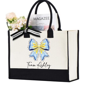 May include: A white canvas tote bag with black trim and handles. The bag has a blue and yellow bow with a white background and the text "Team Ashley" printed on the front.