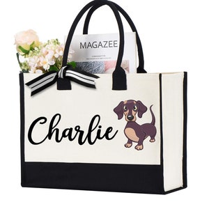 May include: A white canvas tote bag with black trim and handles. The bag has a black and white striped ribbon bow and a cartoon dachshund with the name "Charlie" printed on the front.