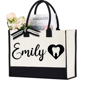 May include: A white canvas tote bag with black trim and handles. The bag has a black and white ribbon bow and the text "Emily" in black cursive lettering. A black heart with a white tooth inside is to the right of the name. The bag is filled with a magazine.