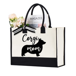 May include: A canvas tote bag with black handles and a black bottom panel. The bag features the words "Corgi mom" with a silhouette of a corgi dog. The bag is filled with flowers and a magazine.