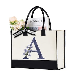 May include: A white canvas tote bag with black trim and a black bottom. The bag has a black and white ribbon tied around the handles. The bag has a large letter "A" printed on the front with a floral design. The bag has the word "MAGAZINEE" printed on a magazine inside the bag.