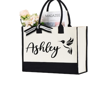 May include: A white canvas tote bag with black trim and handles. The bag has a black hummingbird design and the name "Ashley" printed on the front. The bag is filled with a magazine and flowers.