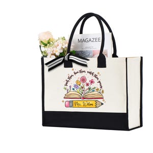 May include: A white canvas tote bag with black trim and handles. The bag has a design of a book with flowers growing out of it and the text "Teach them to love their books, then watch them grow." and "Mrs. Wilson".