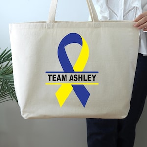May include: A beige canvas tote bag with a blue and yellow ribbon design. The text "TEAM ASHLEY" is printed in black on the bag.