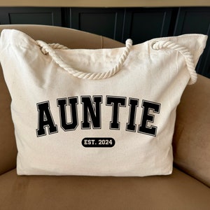 May include: A white canvas tote bag with black rope handles. The bag has the word "AUNTIE" printed in black block letters with a black outline. Below the word is a small rectangle with the text "EST. 2024" printed in black.
