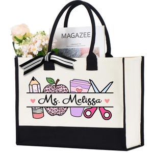 May include: A white canvas tote bag with black trim and handles. The bag features a design of a pencil, apple with leopard print, coffee cup, and scissors. The text "Ms. Melissa" is printed on the bag.