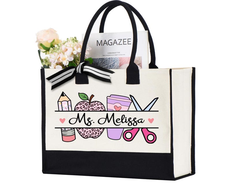 May include: A white canvas tote bag with black trim and handles. The bag features a design of a pencil, apple with leopard print, coffee cup, and scissors. The text "Ms. Melissa" is printed on the bag.