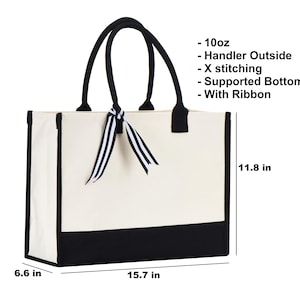 May include: A white canvas tote bag with black trim and a black base. The bag has black handles and a black and white striped ribbon. The bag measures 15.7 inches wide, 11.8 inches tall, and 6.6 inches deep.