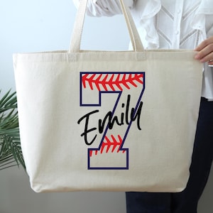 May include: A cream-colored tote bag with a baseball-themed design. The number 7 is outlined in navy blue with red stitching, and the name "Emily" is written in black script. The bag has long handles.
