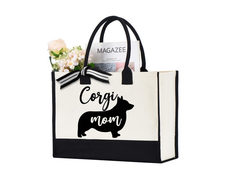 May include: A canvas tote bag with black handles and a black bottom panel. The bag features the words "Corgi mom" with a silhouette of a corgi dog. The bag is filled with flowers and a magazine.