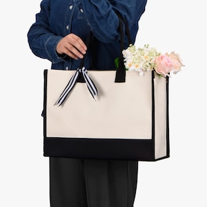 May include: A person is holding a large, white canvas tote bag with black trim. The bag has a black and white striped ribbon tied around the handles. The bag is filled with a bouquet of white and pink flowers.