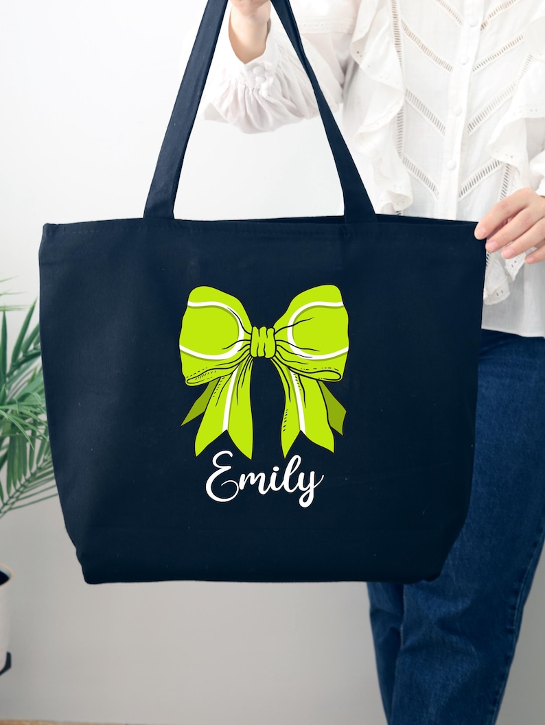 May include: Black canvas tote bag with a green tennis ball bow and the name "Emily" printed in white.