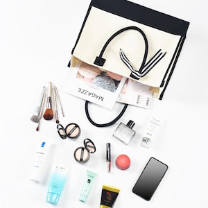 May include: A white and black tote bag with a black ribbon bow. The bag is open and filled with various items, including makeup, skincare products, a magazine, and a mobile phone.