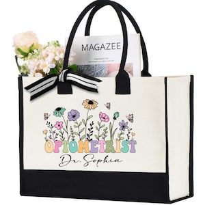 May include: A white canvas tote bag with black handles and a black bottom. The bag features a floral design with the word "OPTOMETRIST" in pastel colors and the name "Dr. Sophia". The bag is filled with flowers and a magazine.