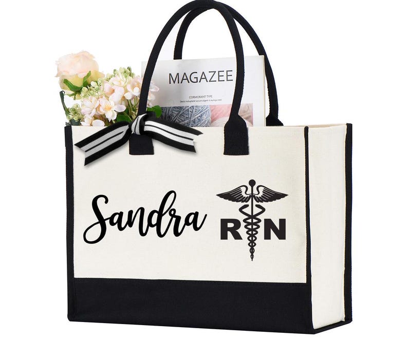 May include: A white canvas tote bag with black trim and handles. The bag has a black and white graphic of a medical caduceus and the text "Sandra RN".