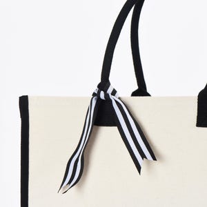 May include: A white canvas tote bag with black trim and black handles. A black and white striped ribbon is tied around the handles.
