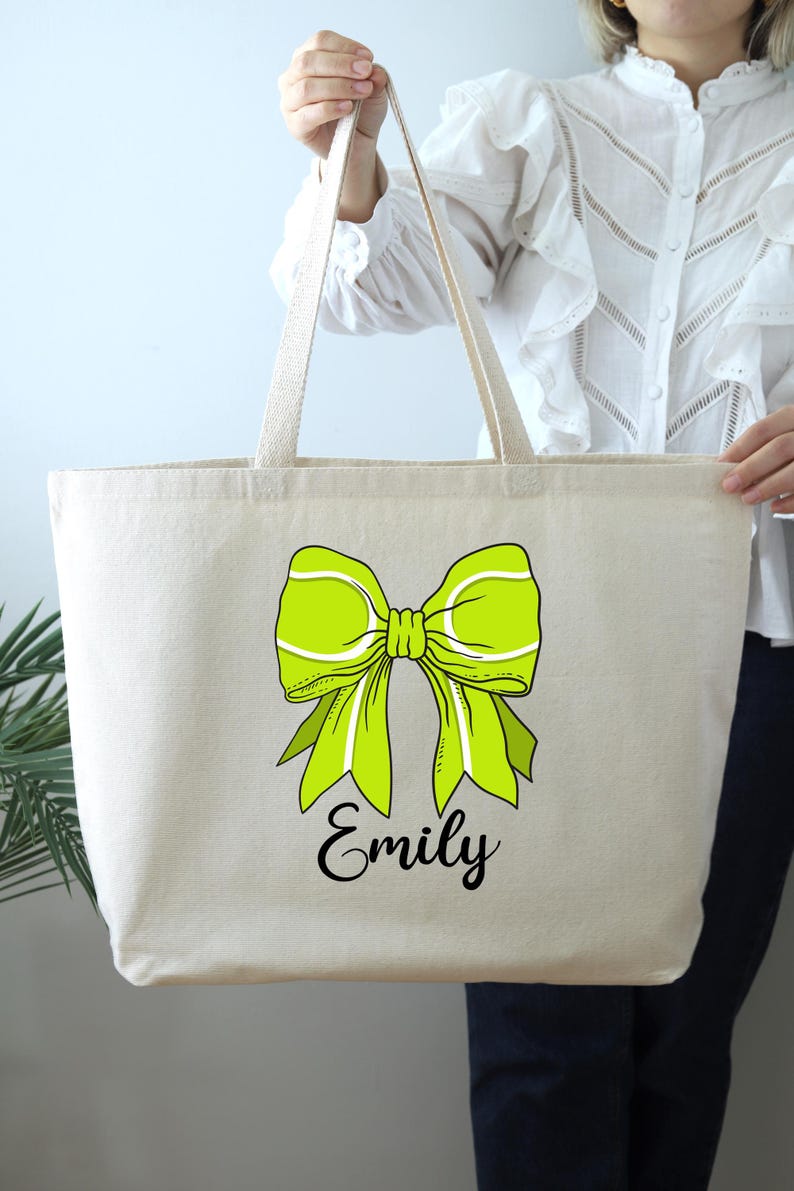 May include: A large, natural-colored canvas tote bag with a green bow and the name "Emily" printed in black.