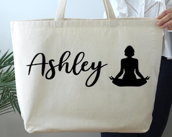 May include: A white canvas tote bag with black handles and the name "Ashley" printed in black cursive script. A black silhouette of a person in a yoga pose is printed on the right side of the bag.