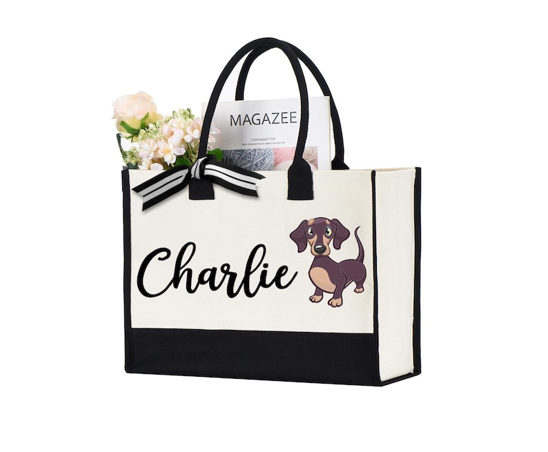 May include: A white canvas tote bag with black trim and handles. The bag has a black and white striped ribbon bow and a cartoon dachshund with the name "Charlie" printed on the front.