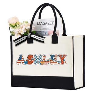 May include: A white tote bag with black handles and a black bottom panel. The name "ASHLEY" is printed on the front in a patriotic font with red, white, and blue patterns. The bag contains flowers and a magazine.