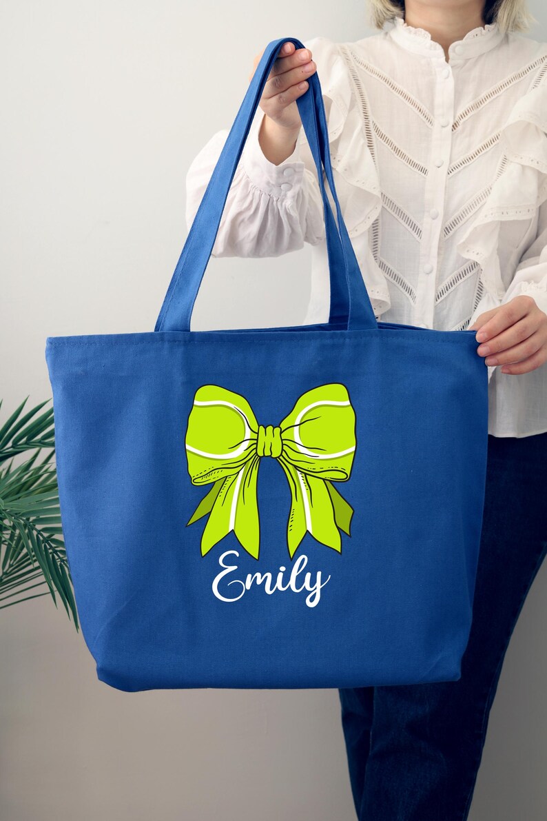 May include: A royal blue tote bag with a green tennis ball bow and the name "Emily" printed in white.