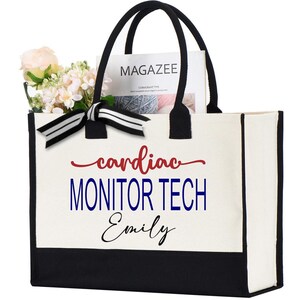 May include: A canvas tote bag with black handles and a black bottom panel. The bag features the text "cardiac MONITOR TECH Emily" in red, blue, and black script. The bag is filled with flowers and a magazine.