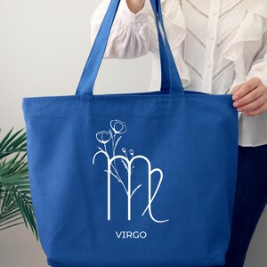 May include: A blue canvas tote bag with a white Virgo zodiac sign and floral design. The text "VIRGO" is printed below the sign.