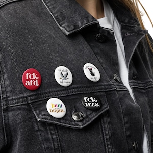 Può includere: Una giacca di jeans nera con cinque diversi bottoni a pressione. I bottoni hanno il seguente testo: "fck afd", "ich fliege auf Magier", "Good Manners are for the Weak", "bunt ist braun" e "fck nzs".
