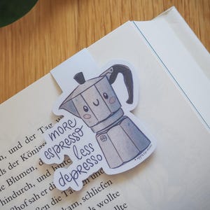 Könnte beinhalten: Ein skurriles Lesezeichen mit einem Espresso-Kocher im Cartoon-Stil und dem Text "more espresso less depresso". Das grau-weiße Lesezeichen ist an der Ecke eines Buches befestigt, mit einer Holzoberfläche im Hintergrund.