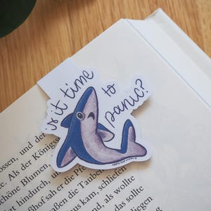 Könnte beinhalten: Ein skurriles Lesezeichen mit einem lächelnden blauen Hai und dem Text "Is it time to panic?" Das Lesezeichen befindet sich auf der Ecke eines Buches mit gedrucktem Text. Das Haidesign hat einen cartoonartigen Stil.
