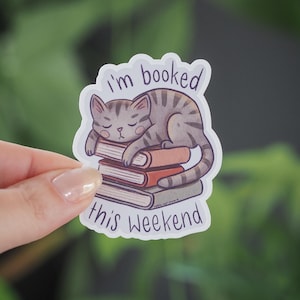 Puede incluir: Una pegatina con un gato atigrado gris durmiendo sobre una pila de libros. La pegatina tiene el texto "I'm booked this weekend". El gato está acurrucado con los ojos cerrados, sugiriendo relajación.