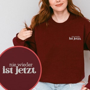 Può includere: Una felpa color borgogna con il testo "nie wieder ist jetzt" in bianco sul davanti.