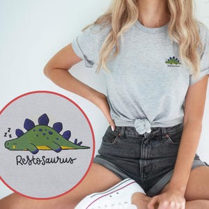 Op de afbeelding: Een grijs gemêleerd t-shirt met ronde hals voorzien van een kleine geborduurde groene dinosaurus met paarse platen op de rug en de tekst Restosaurus. Een cirkelvormige inzet toont een grotere weergave van de slapende dinosaurus.