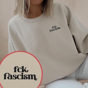 Pode incluir: Camisola bege com a frase "fck fascism" em preto no peito. Um gráfico circular com o mesmo texto também é visível. A camisola é de cor neutra e tem um corte folgado.