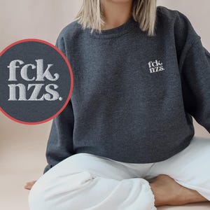 Könnte beinhalten: Ein anthrazitfarbenes Sweatshirt mit Rundhalsausschnitt und weißem Sticktext auf der linken Brust, der fck nzs lautet. Das Kleidungsstück hat lange Ärmel sowie gerippte Bündchen an Ausschnitt, Ärmeln und Saum. Ein runder Ausschnitt zeigt die Stickerei.