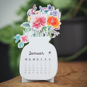 Der Tischkalender im Januar-Blatt auf einem grauen Standfuß vor verschwommenem Grün.