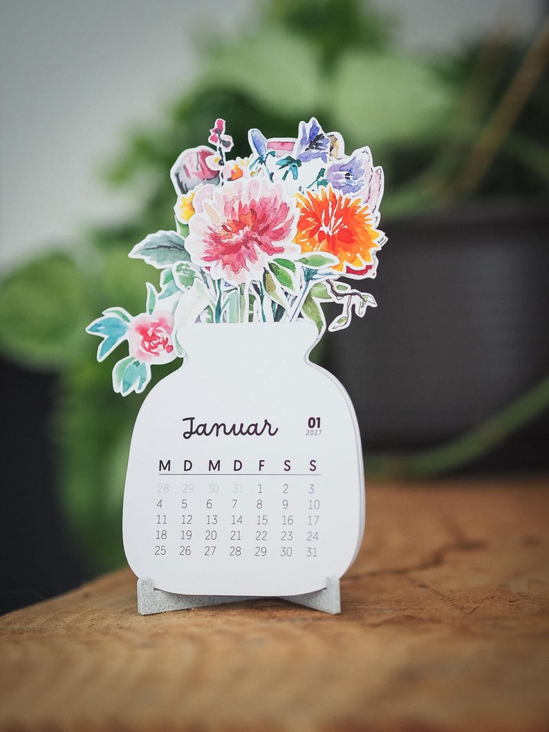 Der Tischkalender im Januar-Blatt auf einem grauen Standfuß vor verschwommenem Grün.