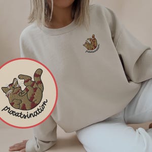 Könnte beinhalten: Ein hellbeiger Pullover mit einem gestickten Katzenmotiv in Rot und Braun und dem Text "procrastination".