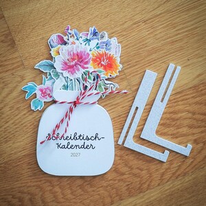 Der geschlossene Tischkalender mit Blumenmotiv und rot-weißer Kordel liegt neben zwei weißen Ständern aus Kunststoff auf einer Holzoberfläche.