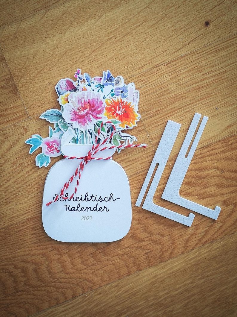 Der geschlossene Tischkalender mit Blumenmotiv und rot-weißer Kordel liegt neben zwei weißen Ständern aus Kunststoff auf einer Holzoberfläche.