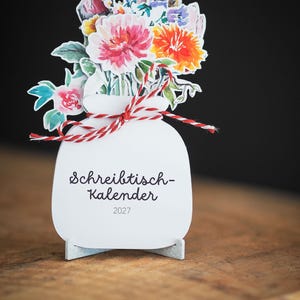 Ein stehender Tischkalender in Vasenform mit einem farbenfrohen Aquarellblumenstrauß und einer rot-weißen Kordel auf einem Holzuntergrund.