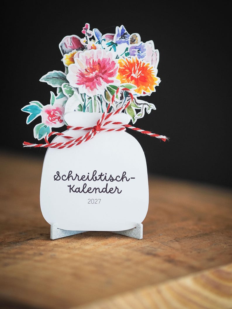 Ein stehender Tischkalender in Vasenform mit einem farbenfrohen Aquarellblumenstrauß und einer rot-weißen Kordel auf einem Holzuntergrund.