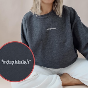 Könnte beinhalten: Dunkelgraues Sweatshirt mit dem Wort "overthinker" in Weiß auf der Brust gestickt. Das Sweatshirt ist im Rundhals-Stil gehalten und scheint aus einem weichen Material zu sein. Das Bild zeigt auch eine Nahaufnahme des gestickten Textes.