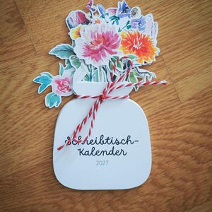 Ein gestapelter Tischkalender 2027 in Vasenform mit einem bunten Aquarell-Blumenstrauß, zusammengehalten durch eine rot-weiße Kordel auf Holztisch.