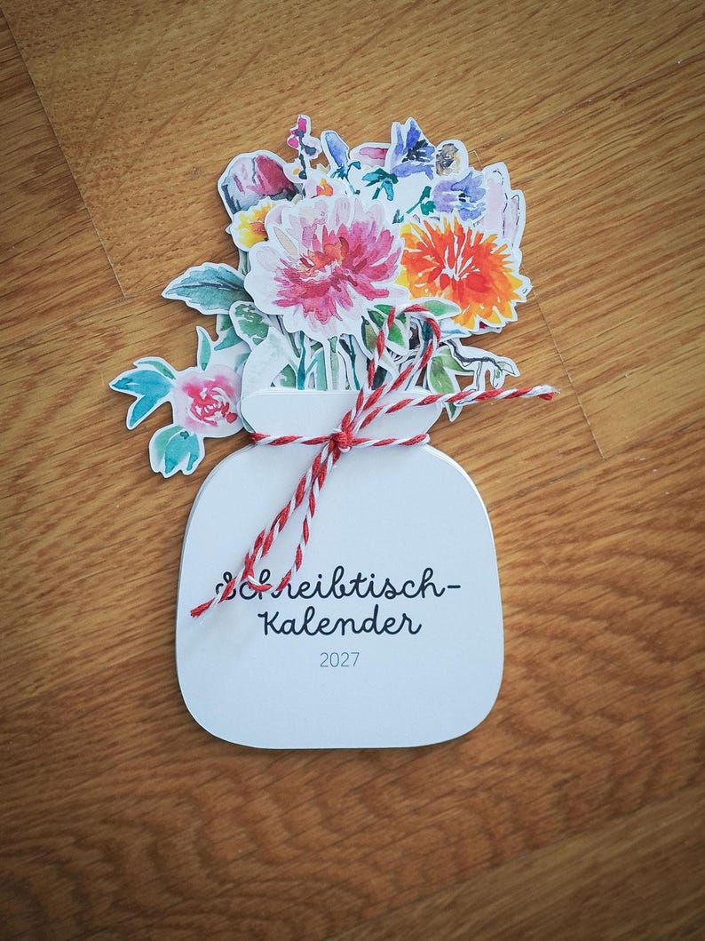 Ein gestapelter Tischkalender 2027 in Vasenform mit einem bunten Aquarell-Blumenstrauß, zusammengehalten durch eine rot-weiße Kordel auf Holztisch.