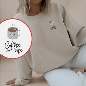 Op de afbeelding: Een beige sweatshirt met een geborduurd koffiekopje en de tekst "Coffee is life".